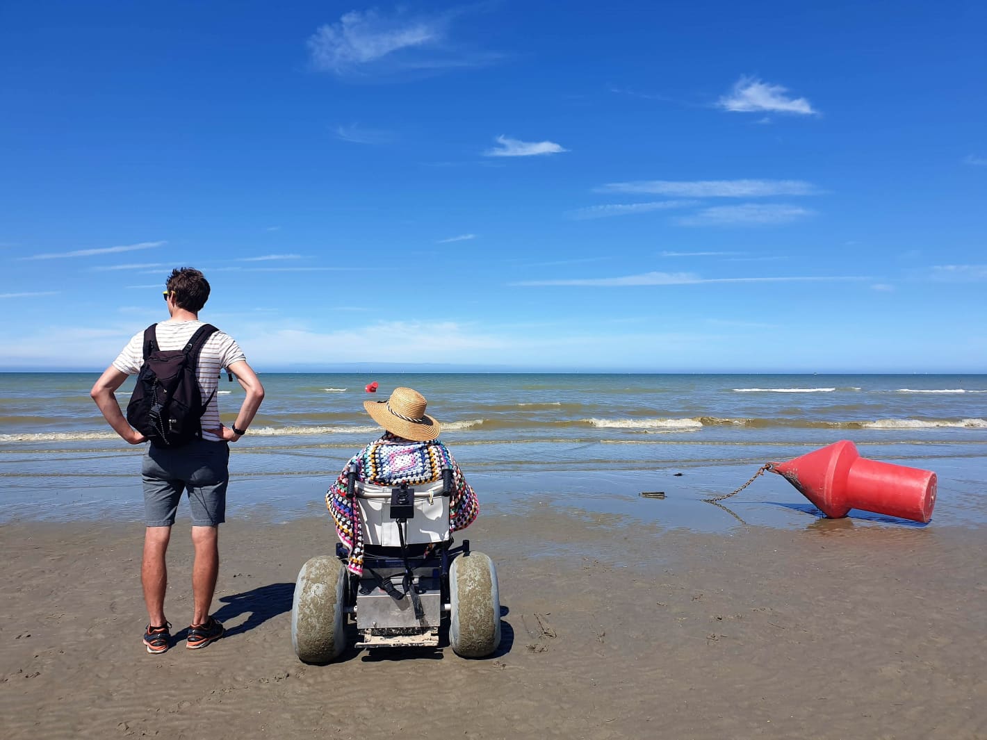 Twee personen op het strand: één staat. De andere zit in een aangepaste rolstoel met dikke banden.
