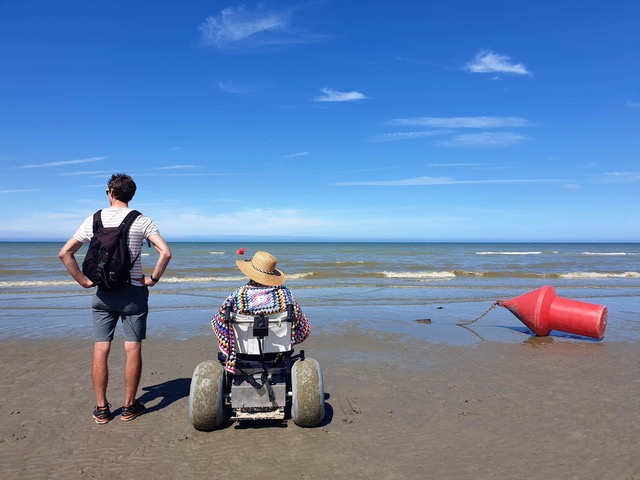 Foto van twee mensen aan de zee op het strand waarvan 1 persoon in een rolstoel