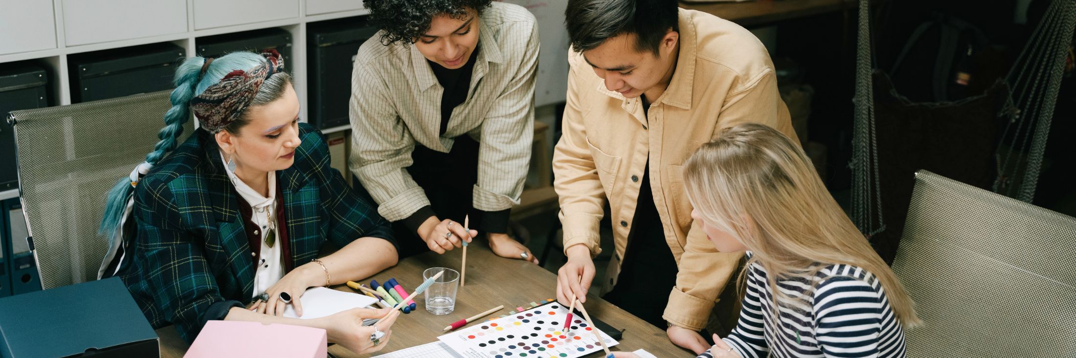 Vier mensen zitten rond een tafel en werken samen aan een project. Eén persoon wijst naar een kleurstaal, terwijl de anderen aandachtig toekijken en deelnemen aan de discussie. De tafel is bedekt met kleurstalen, stiften en andere werkmaterialen. De groep lijkt bezig met een creatief en collaboratief proces.