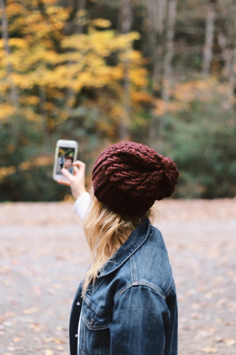 achteraanzicht van jonge blonde vrouw die selfie neemt. bosrijke omgeving. vrouw heeft bordeaux muts aan en jeans vest. Herfst.