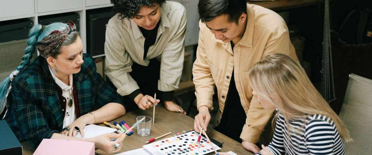 Vier mensen zitten rond een tafel en werken samen aan een project. Eén persoon wijst naar een kleurstaal, terwijl de anderen aandachtig toekijken en deelnemen aan de discussie. De tafel is bedekt met kleurstalen, stiften en andere werkmaterialen. De groep lijkt bezig met een creatief en collaboratief proces.