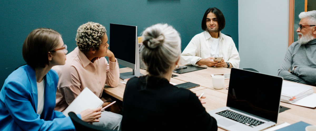 Groep mensen in een kamer die samen een gesprek voeren. Er staan enkele laptops op tafel en er liggen notitieboekjes.